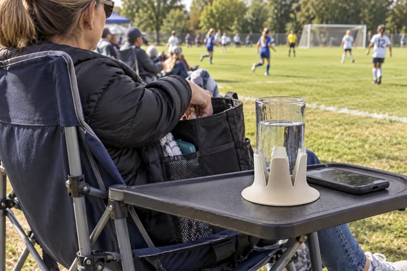 Adult spectator in folding sports chair at local field with open reusable plastic water cup in Steadi on wide level chair-mounted side tray properly supported and fully supporting base. AI illustration.